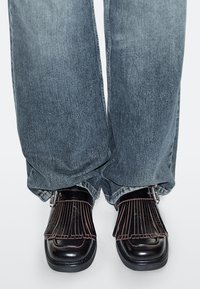 Zwarte leren schoenen met roze franjes, gedragen bij wijd uitlopende blauwe spijkerbroeken. De schoenen hebben een dikke zool en een gesp als accent.