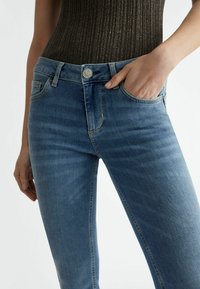 Smala blå denimjeans med en blekt tvätt, fem-ficksdesign och en silverfärgad metallknapp i midjan. Mjuk textur.