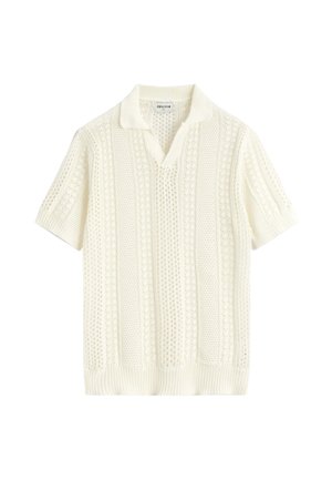 Polo en maille crème à manches courtes avec motifs ajourés texturés et col et bas côtelés, posé à plat sur fond blanc.