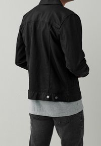 Chaqueta de mezclilla negra con cuello, puños abotonados y dos cierres a presión en la cintura. Usada sobre una camisa gris en capas y jeans oscuros.