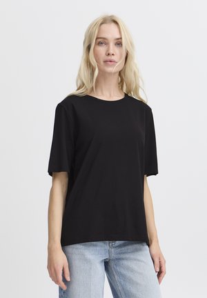 Femme blonde portant un t-shirt noir uni à manches courtes et un jean bleu clair, debout devant un fond blanc.