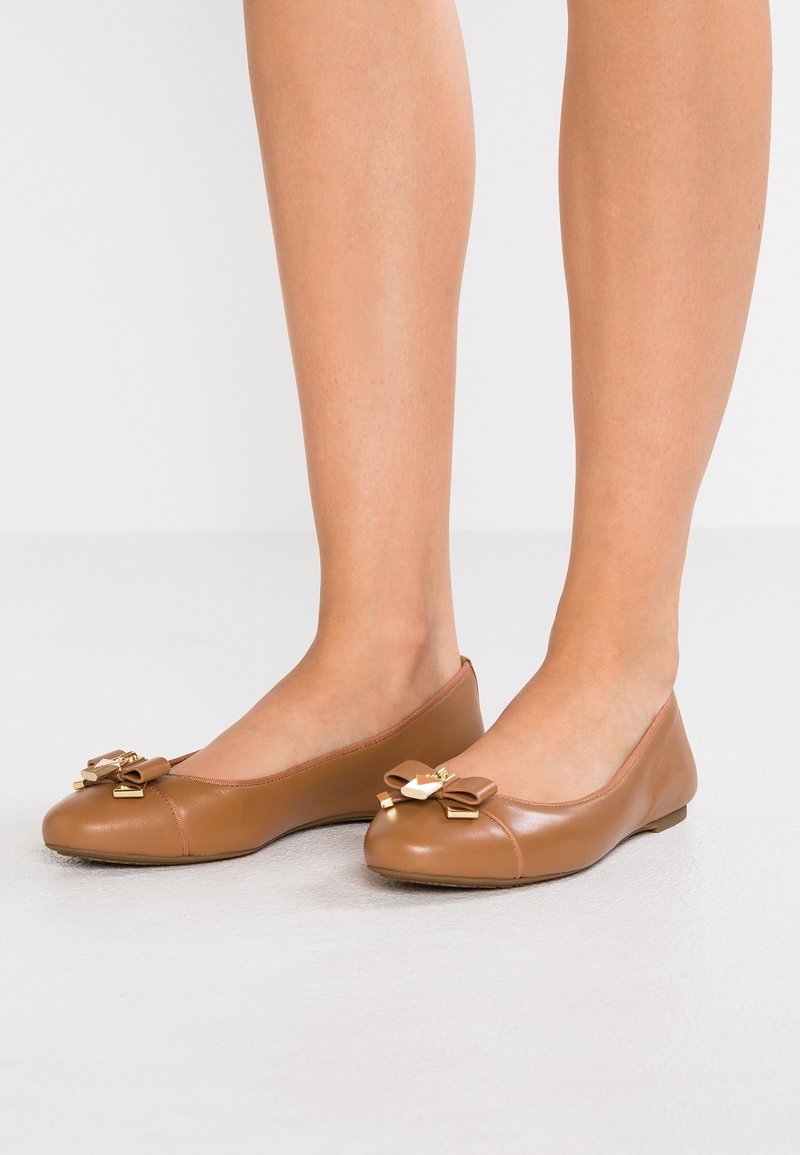 Jambes portant des ballerines en cuir marron clair avec des nœuds décoratifs et des accents dorés sur un fond clair uni.