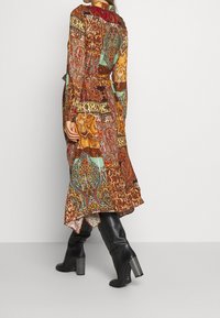 Longue robe multicolore présentant des motifs complexes en rouge, or et bleu sarcelle. Portée avec des bottes noires à talons montant jusqu'aux genoux, créant un look en couches.