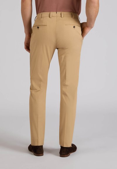 Homme portant un pantalon beige ajusté avec des poches arrière à boutons et des chaussures marron, debout devant un fond clair uni.