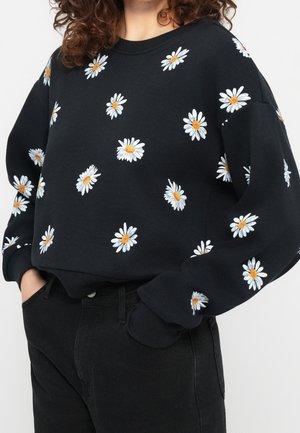 Personne portant un sweat-shirt noir avec des motifs de marguerites blanches et jaunes et un pantalon noir, les mains dans les poches.