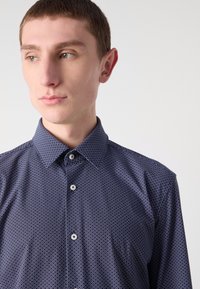 Camicia abbottonata blu navy con un motivo geometrico bianco, colletto strutturato e chiusura frontale con piccoli bottoni in plastica.