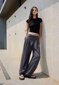 Schwarzes Kurzarmoberteil kombiniert mit grauen weiten Hosen mit Falten und Knopfverschlüssen. Das Modell trägt schwarze Flip-Flops.