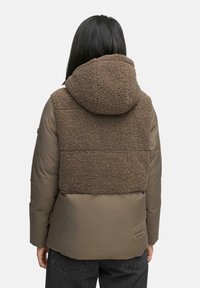 Bruine pufferjack met een fuzzy, gestructureerde bruine fleece achterkant en capuchon, matte afwerking en zijzakken, in combinatie met donkere spijkerbroeken.