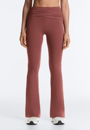 Donna che indossa leggings a vita alta color rosa polveroso a zampa, abbinati a scarpe da ginnastica bianche, in piedi davanti a uno sfondo bianco.