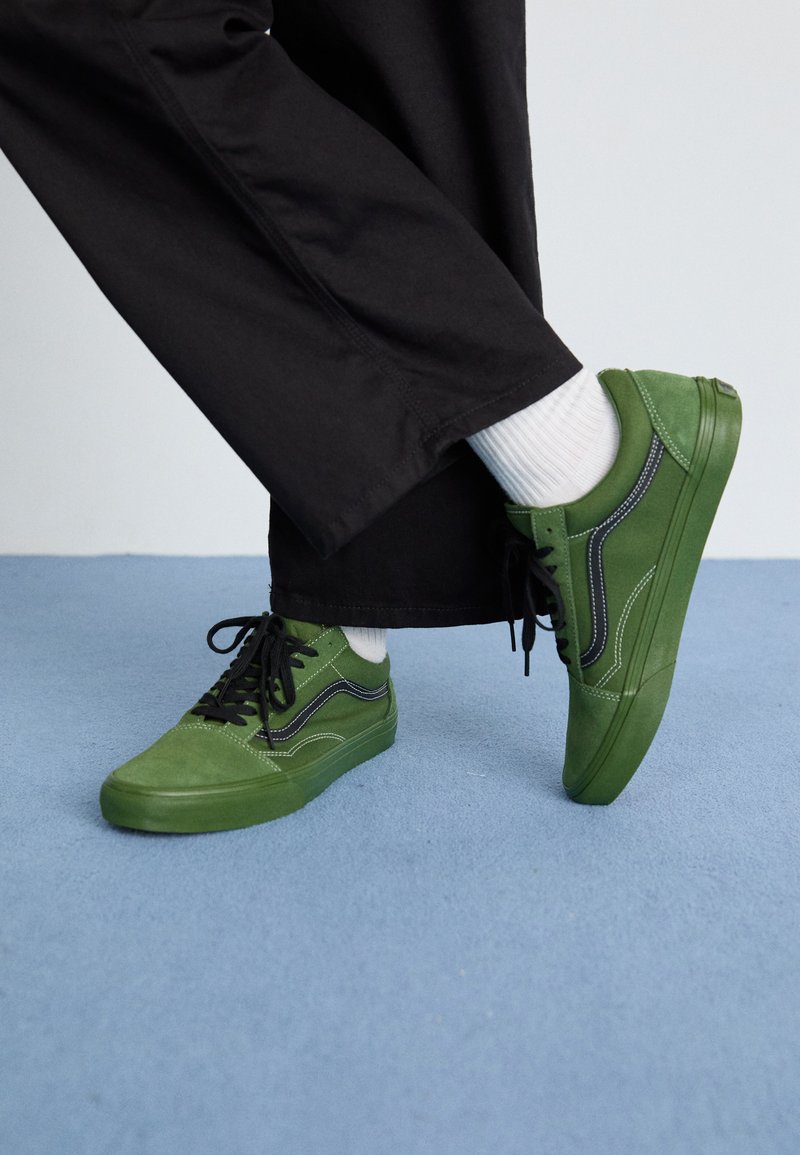 Zapatillas de ante verde con cordones negros, que presentan costuras blancas y un diseño clásico de bajo, combinadas con calcetines blancos y pantalones negros.