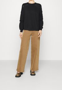 Suéter negro con detalles decorativos de tachuelas en los hombros, combinado con pantalones de pierna ancha en marrón claro y zapatos metálicos tipo slip-on.