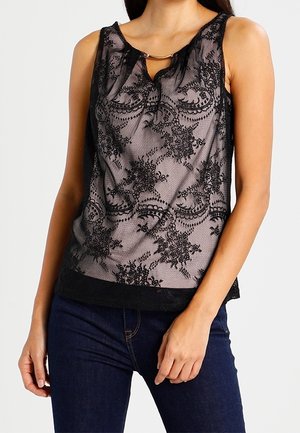 Ærmeløs sort blonde top med blomsterdekorationer båret over et hudfarvet underlag kombineret med mørkeblå jeans på en stående kvinde.