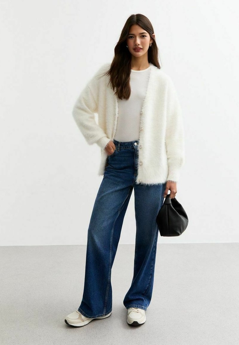 Cárdigan blanco esponjoso con botones en el frente, combinado con jeans de pierna ancha azules y zapatillas crema. La modelo sostiene un bolso negro.