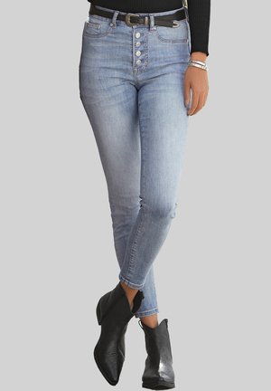 Jeans ajustados de cintura alta en azul claro con cierre de botones en la parte frontal, cinco bolsillos y un ligero desgaste, combinados con botas negras hasta el tobillo.