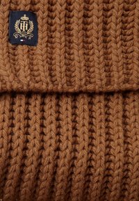 Tissu en maille côtelée épaisse marron avec un bord replié et une étiquette marine ornée d'un emblème brodé en or et d'un petit détail de drapeau.