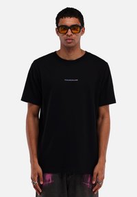Black cotton t-shirt with a crew neckline, featuring a small white logo "TOMORROWLAND" on the chest. Worn with gray jeans showing pink accents.