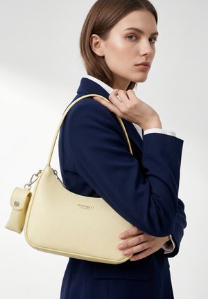 Jonge vrouw in marineblazer die een lichtgele Diana&Co Firenze-handtas over haar schouder draagt tegen een effen achtergrond.