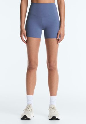 Kobieta w niebieskich sportowych szortach z wysokim stanem, białych skarpetach do połowy łydki i beżowych butach do biegania, stojąca na jednolitym tle.