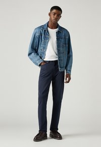 Jeansjacka med ljusblå tvätt, vit T-shirt, marinblå byxor och bruna skor. Jackan har knappar och fickor; byxorna är tapered.