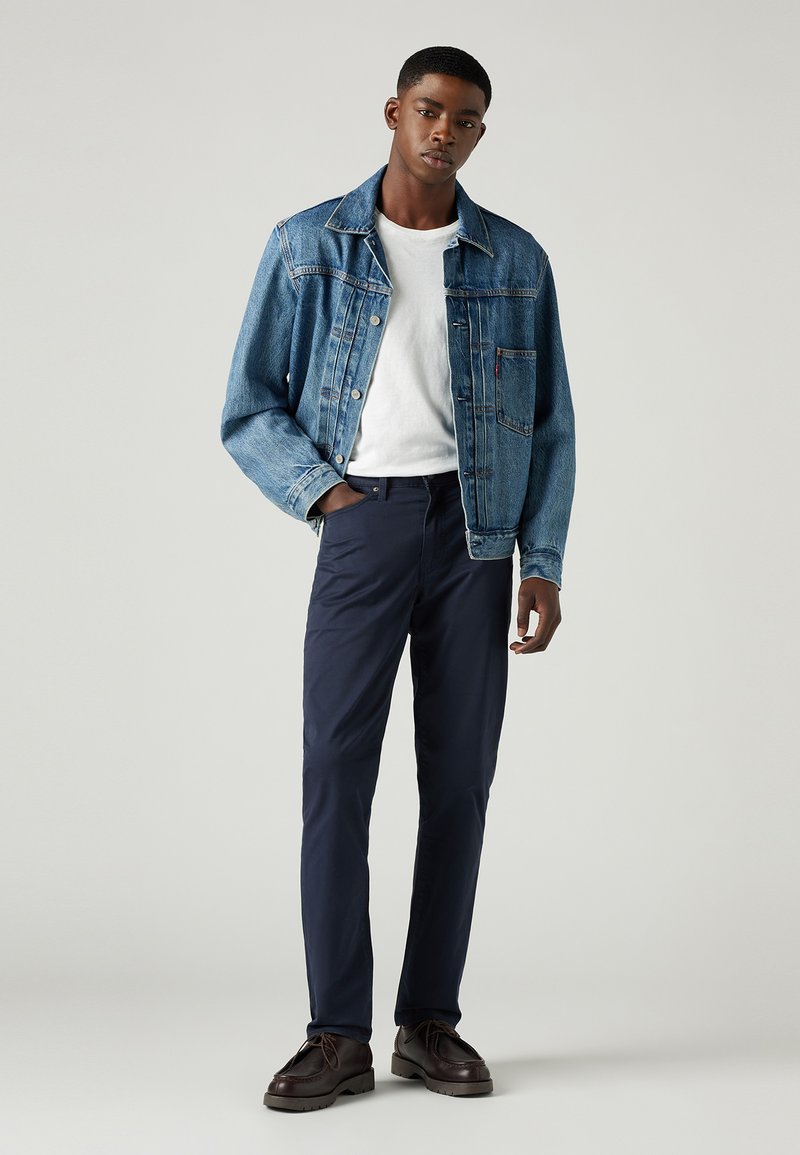Chaqueta de denim con lavado azul claro, camiseta blanca, pantalones navy y zapatos marrones. La chaqueta tiene botones y bolsillos; los pantalones son ajustados.