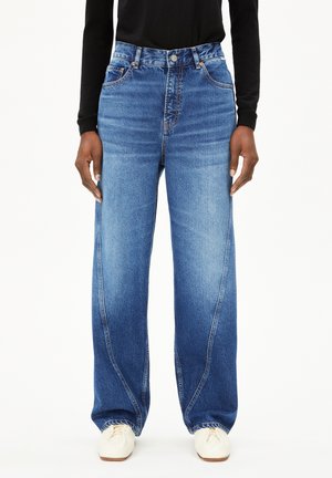Weite blaue Jeans mit mittlerer Taille, leichter Verwaschung und dezenten Details an den Vorderentaschen. Kombiniert mit weißen Schuhen.