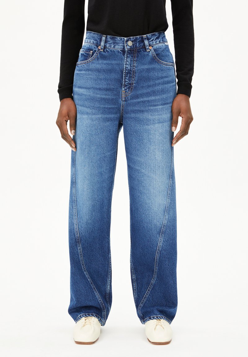 Brede, blauwe denim jeans met een middelhoge taille, lichte vervaging en subtiele details op de voorzakken. Gecombineerd met witte schoenen.