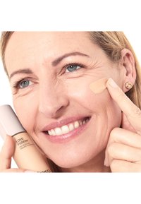 Femme d'âge moyen souriant en appliquant un fond de teint liquide beige sur la joue, tenant le flacon de fond de teint près de son visage.