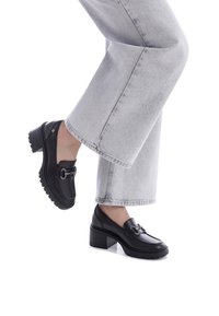 Mocassini in pelle nera con tacco spesso e suola in gomma, dotati di una fibbia metallica decorativa. Indossati con jeans a gamba larga grigi chiari.