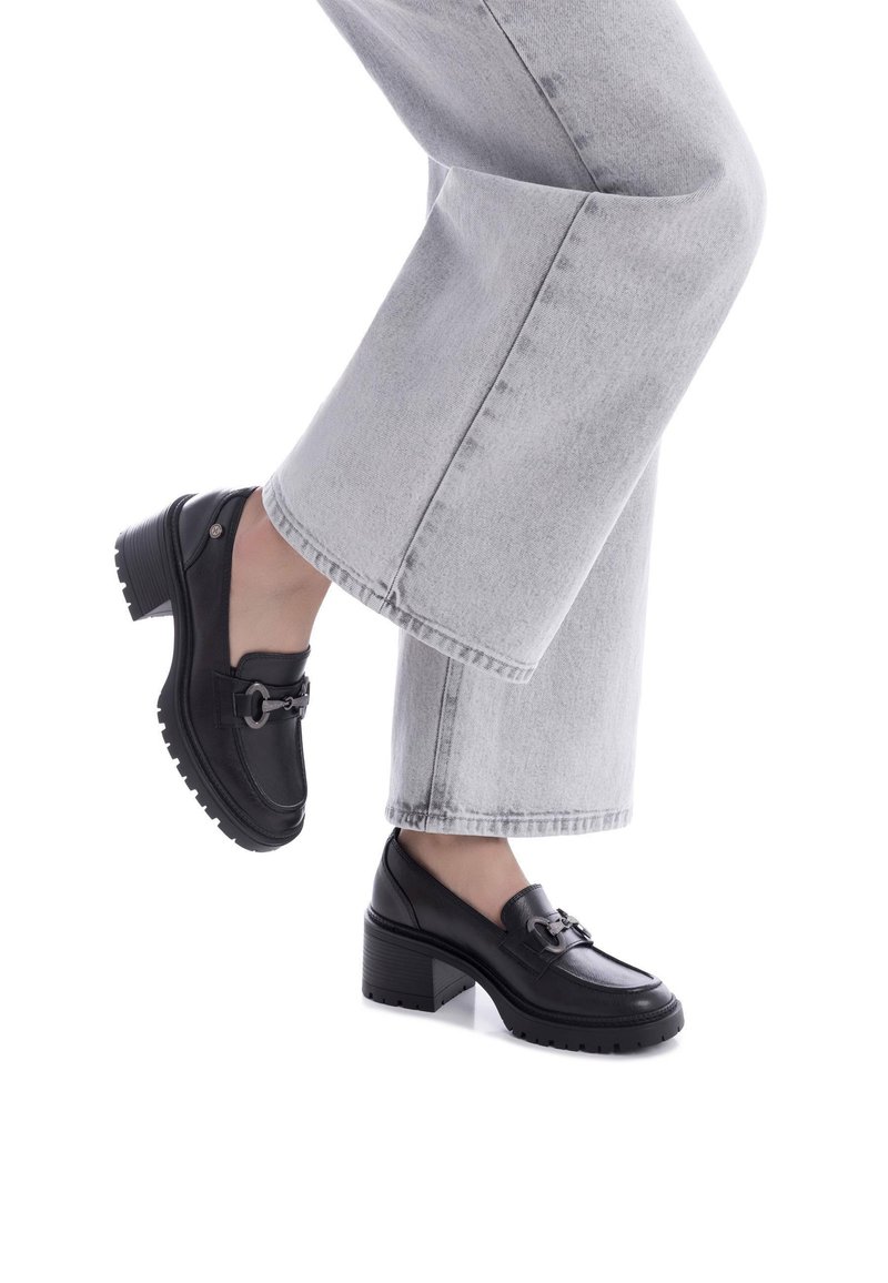 Mocassini in pelle nera con tacco spesso e suola in gomma, dotati di una fibbia metallica decorativa. Indossati con jeans a gamba larga grigi chiari.