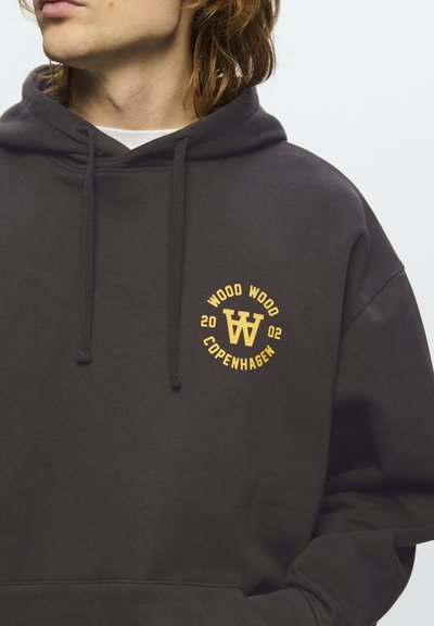 Personne portant un hoodie marron foncé avec un logo jaune "Wood Wood Copenhagen 2002" sur la poitrine, regardant vers la gauche, sur un fond clair.