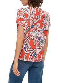 Rot gemusterte Bluse mit kurzen Ärmeln, abstrakten weißen und blauen Designs und einem lockeren Schnitt, kombiniert mit blauen Jeans.