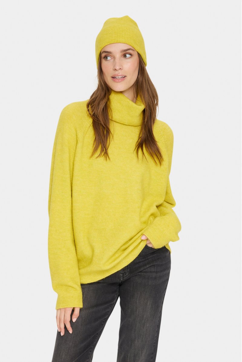 Keltainen oversized-sweateri, jossa on korkea cowl-kaulus ja ribbitetyöt hihansuissa, yhdistettynä vastaavaan ribbitettyyn pipoon ja tummiin farkkuihin. Sujuva pinta.
