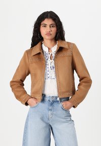 Brun kunstlærjakke med strukturert krage, sølvnagler på sømmene og glidelås foran. Båret over en hvit bluse og denimjeans.
