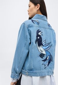 Γυναίκα που φοράει ανοιχτό μπλε τζιν μπουφάν με μεγάλο κεντημένο σχέδιο μαύρου και μπλε πουλιού στην πλάτη, στραμμένη ελαφρώς πλάγια.
