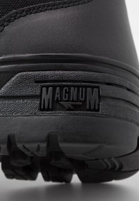 Svart sko med en strukturerad gummisula och en framträdande "MAGNUM"-logotyp. Hållbart material med en robust design och en lågprofil-silhuett.