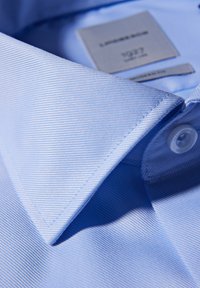 Lichtblauwe shirtkraag met diagonale ribbels, voorzien van een witte knoop en een verzorgingslabel aan de binnenkant, die de strakke lijnen en stofdetails benadrukt.
