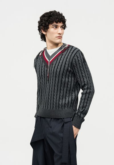 Jeune homme aux cheveux bouclés foncés portant un pull en maille torsadée gris et noir à rayures avec des accents rouges et un pantalon ample à fines rayures foncées, regardant vers la droite.