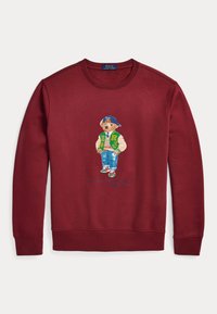 Sudadera burdeos con un oso bordado que lleva una chaqueta varsity verde y blanca, jeans azules y una gorra azul. Puños y cinturilla de punto.