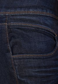 Mörkblå denimjeans har subtil textur, orange sömnad och en framficka. Tyget är något bleknat på vissa ställen.