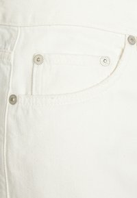 Jeans en denim blanc avec une petite poche avant équipée de rivets métalliques argentés, un tissu texturé et une finition lisse.