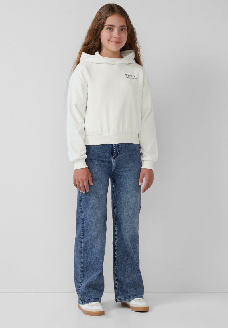 Weiße, gekürzte Kapuzenjacke mit Kordeln, mit dem Schriftzug "Berlin". Kombiniert mit weit geschnittenen blauen Jeans und weißen Sneakern mit beigen Sohlen.