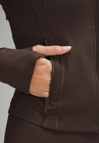 Brun atletisk jakke lavet af glat stof, med en lynlåslomme og tommelhuller i manchetterne, samt flade sømdetaljer.