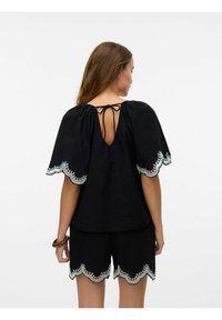 Top in cotone nero con bordi ricamati bianchi a scallop, maniche a palloncino e chiusura con fiocco sul retro, abbinato a pantaloni corti coordinati.