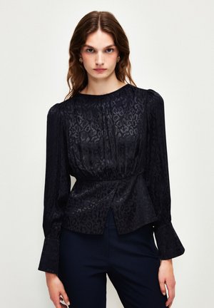 Blouse noire à manches longues avec une texture subtile en imprimé léopard, un col arrondi, une taille froncée et des poignets évasés. Accentuée d'un devant plissé.