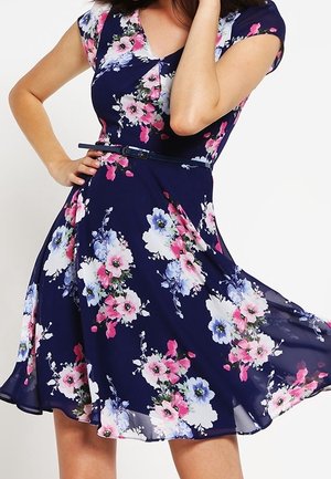 Kobieta w granatowej sukience z krótkim rękawem, zdobionej różowym, białym i fioletowym wzorem kwiatowym oraz cienkim granatowym paskiem, trzymająca sukienkę na bok.