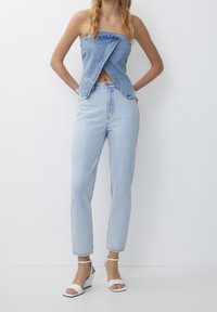 Top sin tirantes de mezclilla con un nudo en el centro, combinado con jeans azul claro de cintura alta y sandalias de cuña blancas. Textura suave. Diseño casual.