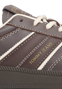 Brązowy sneaker ze skóry z białymi zygzakowatymi paskami, złotym logo "TOMMY JEANS" oraz teksturowaną gumową podeszwą z pionowymi rowkami.