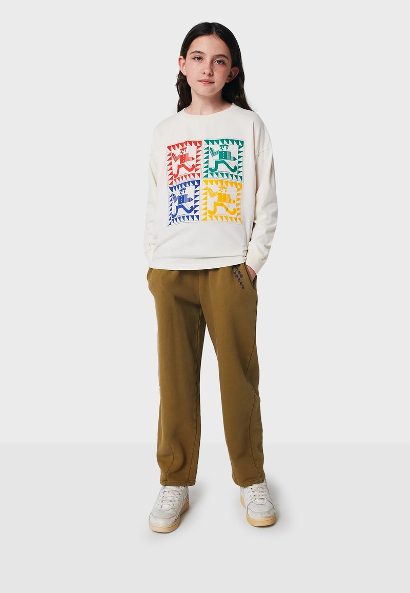 Cremefarbener Sweatshirt mit bunten geometrischen Drucken in Rot, Grün, Blau und Gelb; kombiniert mit olivgrünen Jogginghosen und weißen Sneakers.