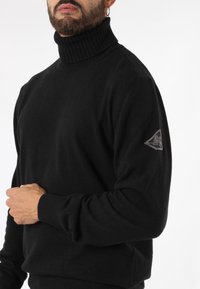 Maglione nero a collo alto con colletto e polsini a costine. Presenta una patch con logo sulla manica sinistra. Realizzato in un morbido tessuto testurizzato.