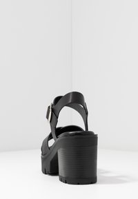 Sandales à talons hauts noires avec une plateforme épaisse, une sangle ajustable au niveau de la cheville et en cuir lisse. Semelle extérieure en caoutchouc texturé.